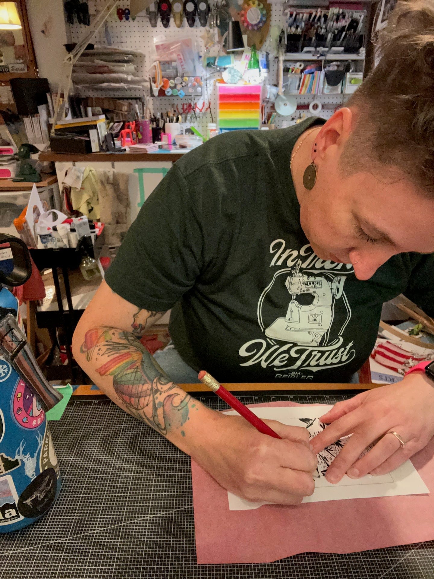 Person with tattoos working at a craft desk, writing on a piece of paper.